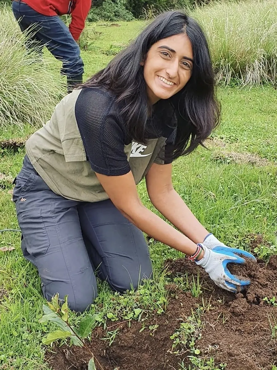 Ashni kneeling on grass in gardening gloves, smiling as she presses soil around a newly planted sapling.