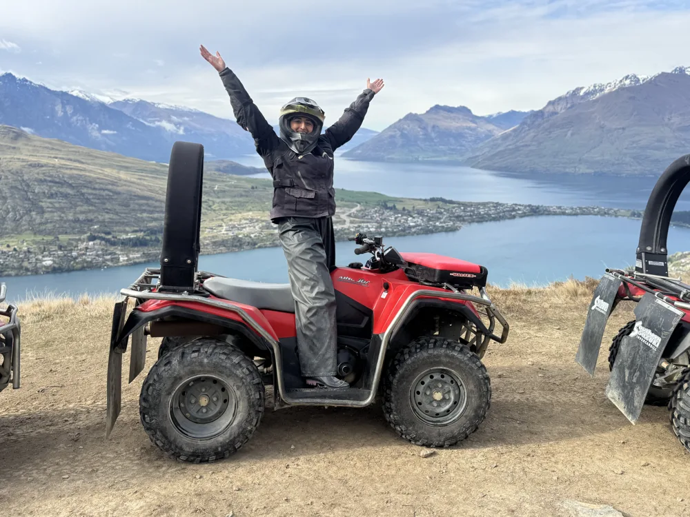 Ashni in a helmet and waterproofs standing on a red quad bike with both arms raised, above a blue lake and mountains near Queenstown.